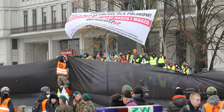Mężczyzna w kamizelce z logiem „Straż Marszu Niepodległości” mocuje czarne płachty, które mają zasłonić pikietę „Obywatele RP”.
